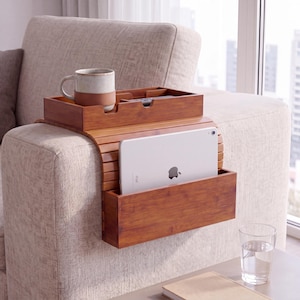 May include: A brown wooden sofa armrest tray with a flexible slatted design. It features a top compartment for a mug and remote, and a front-facing pocket holding a silver tablet. The tray rests securely over the arm of a beige upholstered sofa.