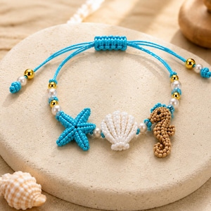 Op de afbeelding: Een kralenarmband met een blauwe zeester, een witte schelp en een beige zeepaardje bedel. De armband is gemaakt van felblauw koord, witte parels en goudkleurige kralen, afgewerkt met een verstelbare schuifknoopsluiting.