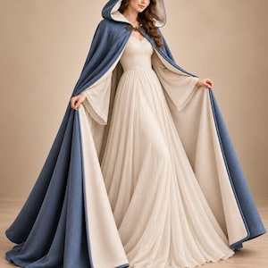May include: A floor-length hooded cloak in a dusty blue hue with a cream-colored lining, featuring a decorative bronze-toned metal clasp at the neckline. The cloak is worn over a long, flowing cream-colored dress with sheer, wide sleeves.