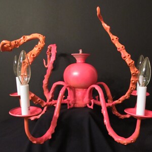 May include: A pink octopus-shaped chandelier with eight arms and six light bulbs. The chandelier is made of metal and has a pink finish.