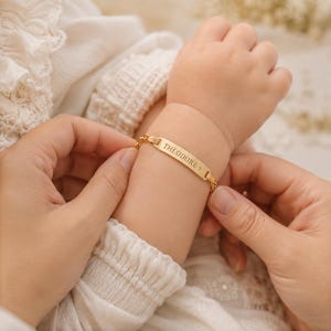 May include: A gold-toned metal identification bracelet featuring a rectangular center plate engraved with the name THEODORE followed by a small cross symbol. The plate is attached to a delicate gold-toned chain link band worn around a wrist.