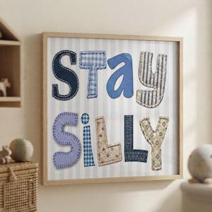 Op de afbeelding: Een vierkant ingelijst wandkunstwerk met de tekst Stay Silly. De letters zijn ontworpen met diverse blauwe en beige stoffen patronen, waaronder denim, gingham en sterrenprints, tegen een achtergrond met lichtblauwe en witte verticale strepen.