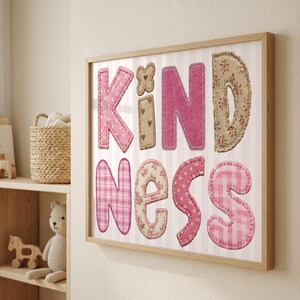Op de afbeelding: Een ingelijst wandkunstwerk met het woord KINDNESS in grote, stofachtige letters. Elke letter heeft een uniek patroon, waaronder roze gingham, bloemenprints en stippen, geplaatst tegen een lichtroze en wit gestreepte achtergrond.