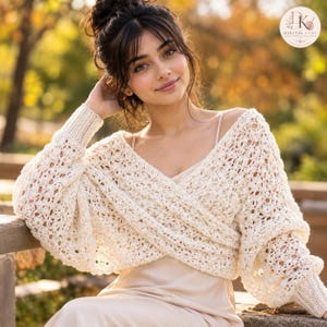 May include: A cream-colored, long-sleeved crochet shrug featuring an openwork, textured pattern with a wrap-style front and ribbed cuffs. The garment is worn over a simple, sleeveless, light-colored slip dress with thin spaghetti straps.
