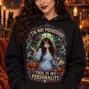 Puede incluir: Una sudadera negra con capucha que presenta un estampado gráfico de una persona con vestido azul rodeada de objetos flotantes, calabazas y humo verde. El texto dice I'M NOT POSSESSED. THIS IS MY PERSONALITY. El diseño tiene un borde morado.