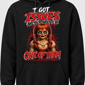 Puede incluir: Una sudadera con capucha negra que presenta un estampado gr&aacute;fico de una mu&ntilde;eca con trenzas rojas. El texto I GOT ISSUES AND YOU'RE ABOUT TO BE ONE OF THEM aparece en letras rojas y blancas, rodeado de salpicaduras rojas en el frente.