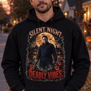 Puede incluir: Una sudadera negra con capucha que presenta un estampado gráfico de una figura enmascarada sosteniendo un cuchillo frente a una casa y una luna llena. El texto SILENT NIGHT aparece arriba y el texto DEADLY VIBES está impreso en rojo debajo.