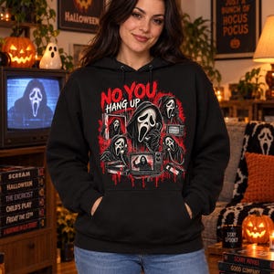 Puede incluir: Una sudadera negra con capucha que presenta un estampado gráfico con el texto NO YOU HANG UP en rojo. El diseño incluye varias ilustraciones de una figura enmascarada sosteniendo un teléfono, rodeada de salpicaduras de pintura roja y televisores.