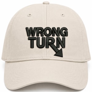 May include: A beige baseball cap featuring the black embroidered text WRONG TURN on the front. The word TURN includes a downward-pointing arrow integrated into the letter N. The cap has a structured crown, a curved brim, and stitched eyelets.