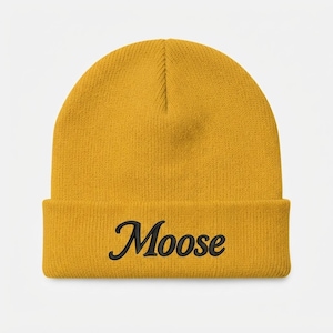Op de afbeelding: Een mosterdgele muts met omslag waarop het woord Moose in zwarte cursieve letters op de voorzijde is geborduurd. De gebreide muts heeft een ronde bovenkant en een dikke geribbelde textuur over de gehele stoffen constructie.