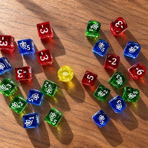May include: A collection of translucent six-sided dice in red, blue, green, and yellow colors. Each die features white printed numbers and shield icons with positive or negative values. One yellow multi-sided die is positioned in the center.