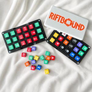 Puede incluir: Un juego Riftbound que incluye dos bandejas rectangulares negras llenas de pequeñas fichas cuadradas en rojo, verde, amarillo, naranja, azul, morado y negro. Las fichas muestran varios símbolos y números. Se incluye una tapa blanca con el texto naranja Riftbound.
