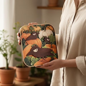 May include: A square wooden-framed wall art piece featuring a layered paper cutout design of dark brown crows perched among orange and white pumpkins with green foliage. The artwork has rounded corners and two small metal loops at the top for hanging.
