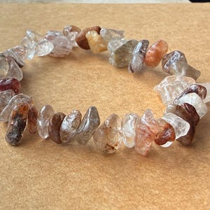 Pode incluir: Uma pulseira elástica composta por fragmentos irregulares de pedras preciosas polidas em tons de transparente, branco, bege e castanho-avermelhado. As pedras estão enfiadas num círculo, exibindo variações naturais de cor e textura.