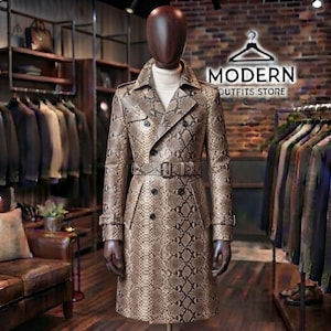 May include: A double-breasted trench coat featuring a brown and tan snakeskin pattern, a notched lapel, and a matching belt with a buckle. The coat is displayed on a dark brown mannequin torso in a retail setting with clothing racks in the background.