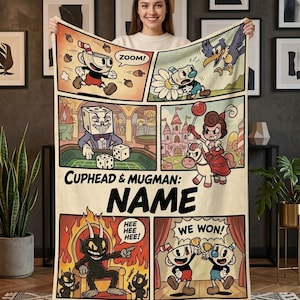 May include: A cream-colored fleece blanket featuring a multi-panel comic strip design with Cuphead and Mugman characters. The print includes the text "CUPHEAD & MUGMAN: NAME", "ZOOM!", "HEE HEE HEE!", and "WE WON!" alongside various illustrated scenes.