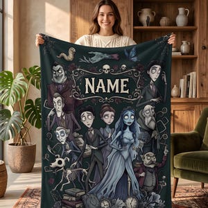 May include: A dark-themed fleece blanket featuring a gothic-style illustration of various animated characters, including a bride and groom, a skeleton dog, and a skull, with the word NAME centered in a decorative frame at the top of the design.