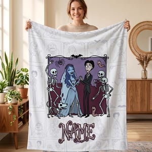 May include: A white fleece blanket featuring a central purple-bordered illustration of a bride and groom with skeletons and a dog. The design includes the text NAME in a stylized font, surrounded by a repeating light gray outline pattern of characters.