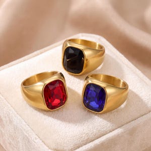 May include: Three gold-toned rings featuring large, rectangular, faceted stones. One ring has a deep red stone, one has a dark blue stone, and one has a black stone. Each ring has a smooth, polished gold-colored band and a wide setting.