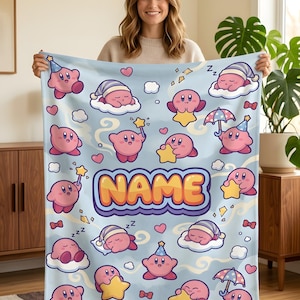 May include: A light blue fleece blanket featuring a repeating pattern of pink Kirby characters in various poses, including sleeping on clouds, holding yellow stars, and wearing party hats. The word NAME is printed in large orange letters.