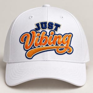 Könnte beinhalten: Eine weiße Baseballkappe mit dem gestickten Text "JUST VIBING" auf der Vorderseite. Das Wort "JUST" ist in dunkelblauen Blockbuchstaben gehalten, während "Vibing" in einer großen, orangefarbenen Schreibschrift mit dunkelblauem Rand und hellblauem Schatten erscheint.