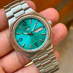 May include: A Seiko 5 automatic watch with a vibrant turquoise sunray dial, silver-tone hands, and hour markers. The watch features a stainless steel case and link bracelet, a day-date window at the three o'clock position, and Arabic numerals at 6, 9, and 12.