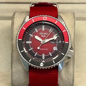Pode incluir: Um relógio de pulso automático Seiko 5 com mostrador vermelho com padrão de raios de sol, ponteiros e marcadores prateados e uma janela de dia e data na posição das três horas. O relógio tem uma luneta rotativa vermelha e preta e uma bracelete de nylon vermelha.