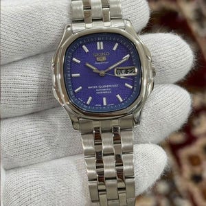 Pode incluir: Um relógio automático Seiko Superior prateado com mostrador roxo texturizado, marcadores de hora prateados e janela de dia e data na posição das três horas. O relógio possui uma pulseira de elos de aço inoxidável e caixa prateada polida.
