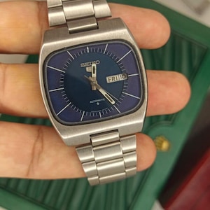 Puede incluir: Un reloj de pulsera automático Seiko 5 con caja cuadrada de acero inoxidable y brazalete de eslabones de metal cepillado a juego. El reloj tiene una esfera azul oscuro con manecillas e índices plateados y una ventana de día y fecha con FRI 10.