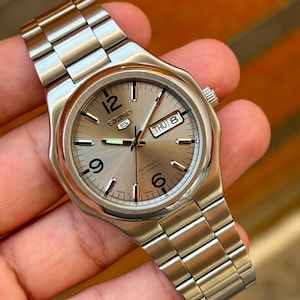 May include: A silver-toned Seiko 5 automatic wristwatch with a metallic champagne-colored dial, featuring Arabic numerals at 6, 9, and 12 o'clock, a day-date window at the 3 o'clock position, luminous hands, and a brushed stainless steel link bracelet.