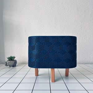 May include: A dark blue cylindrical planter featuring a repeating geometric pattern of embossed triangles and dots. The planter is supported by three light brown, tapered wooden legs that provide elevation and stability to the structure.