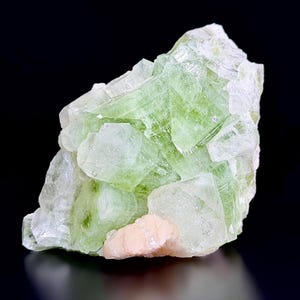May include: A raw mineral specimen featuring translucent, pale green crystalline structures with sharp, geometric facets. A small, opaque, peach-colored mineral deposit is attached to the lower front section of the cluster against a black background.