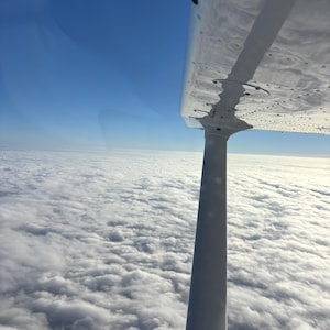 May include: A view from an airplane window showing the metallic white wing and a vertical support strut extending downward. Below the aircraft, a vast expanse of white clouds stretches toward the horizon under a clear, bright blue sky.