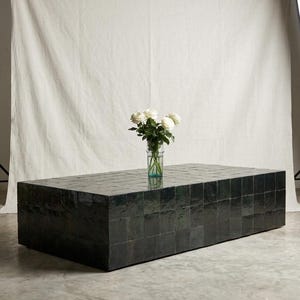 May include: A rectangular coffee table covered in dark green, glossy, square tiles with a smooth, reflective finish. A clear glass vase containing a bouquet of white roses with green stems and leaves sits on the center of the table surface.