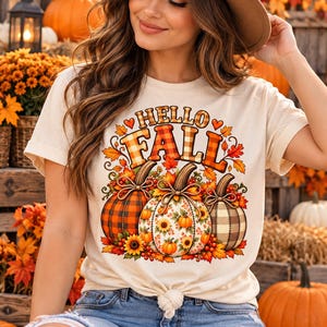 Könnte beinhalten: Ein cremefarbenes Kurzarm-T-Shirt mit einem Grafikdruck, der den Text "HELLO FALL" in orangefarbenen und braunen karierten Buchstaben zeigt. Unter dem Text befinden sich drei Kürbisse mit Karomustern und Blumenmotiven, umgeben von Herbstblättern.