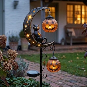 May include: A black metal garden stake featuring a crescent moon with star cutouts, a black cat silhouette, and two hanging orange pumpkin lanterns with carved jack-o'-lantern faces. The structure includes a small solar panel at the base.