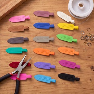 May include: A collection of multi-coloured metal hair clips in various shades including pink, brown, blue, yellow, green, orange, purple, and black, accompanied by a pair of metal pliers, a spool of wire, and several small metal jump rings.