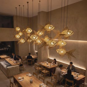 May include: A large installation of multiple hanging pendant lights shaped like fish, crafted from woven light brown bamboo or rattan material. Each fish-shaped light fixture contains an internal bulb, suspended at varying heights above tables.
