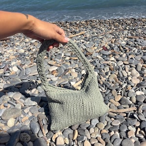 May include: A sage green handbag crafted from thick, chunky knit yarn. The bag features a structured, triangular silhouette with a single integrated handle, showcasing a consistent vertical ribbed texture throughout the entire exterior surface.