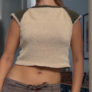 May include: A short-sleeved knit top featuring a cream-colored body and dark olive green raglan sleeves. The garment has a round neckline and a cropped hemline, constructed with a visible stockinette stitch pattern throughout the fabric.