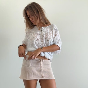 May include: A white short-sleeved blouse featuring a delicate lace panel across the chest and a round neckline, paired with a beige knit mini skirt that has an elasticized waistband, a drawstring tie, and a small patch pocket on the right side.