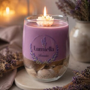 Peut inclure: Une bougie en verre transparent remplie de cire violette, avec une mèche en forme de fleur blanche. Le verre porte l'inscription Lumiella Lavender entourée d'une couronne de lavande. La base contient du sable et divers coquillages.