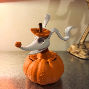 Può includere: Una statuetta di cane fantasma bianco con muso lungo e orecchie cadenti, con una piccola zucca arancione sul naso. Il cane fantasma indossa un cappello a forma di zucca arancione e poggia su una base a forma di zucca arancione testurizzata.