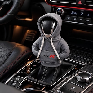 May include: A grey fabric hoodie cover for a car gear shift knob, featuring a front kangaroo pocket with the text Type R printed in black and red. The hoodie includes a hood and white drawstring cords, fitted over a black leather gear shifter.