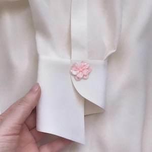 May include: A crisp white long-sleeved shirt featuring a structured cuff. The cuff is fastened with a single, light pink, multi-petaled flower-shaped button that adds a distinct floral detail to the otherwise plain white fabric of the garment.