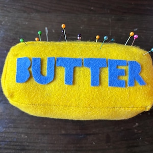 May include: A rectangular yellow felt pincushion shaped like a stick of butter with the word BUTTER appliqued in blue felt letters on the front. Several sewing pins with colorful plastic heads are inserted into the top of the yellow felt.