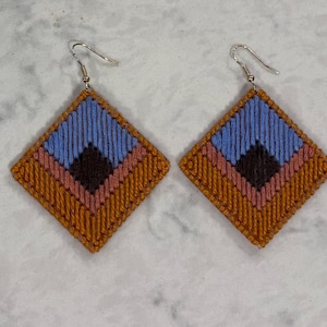 Peut inclure: Une paire de boucles d'oreilles en forme de losange avec une broderie de fil complexe en couches concentriques bleu clair, brun foncé, rose poudré et jaune moutarde. Chaque boucle est suspendue à un crochet en métal argenté.