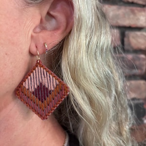 Peut inclure: Une boucle d'oreille en forme de losange avec une broderie complexe dans des tons de rose poudré, bordeaux profond et orange brûlé. Le motif géométrique est réalisé avec des fils superposés sur une base structurée, suspendue à un crochet doré.