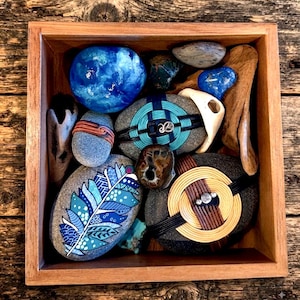 May include: A square wooden box containing a collection of smooth stones, some painted with blue feather patterns, others wrapped in colorful cord, wire, or rattan, alongside pieces of driftwood and small polished blue and brown decorative rocks.