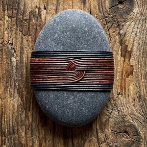 May include: A smooth, oval-shaped gray stone wrapped horizontally around its center with multiple strands of thin, dark black and reddish-brown cord. The cords are tightly wound and intersect in the middle to form a distinct, woven knot pattern.
